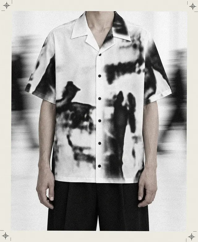 Une personne inventée, coupée au cou, porte la chemise Rift blanche à taches noires floues et se tient immobile dans une salle blanche, pendant qu'autour passent des silhouettes en mouvement.