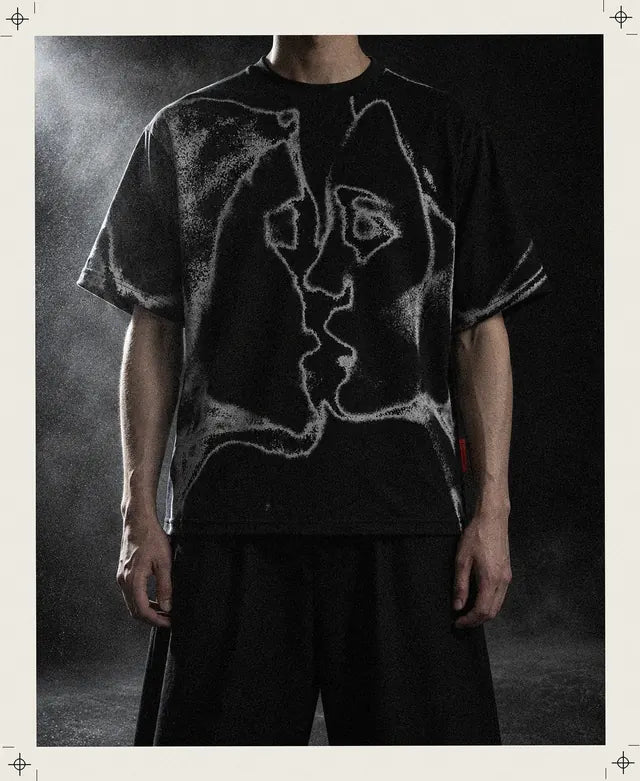 En påhittad person, i bild från halsen och nedåt, bär den svarta T-shirten Unkiss med de två grå profilerna som kysser varandra, i ett svart rum som en tunn dimma drar igenom.