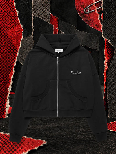 The black Brokenheart zip hoodie in a dark paper room, under a large red paper heart broken in two and held together with a safety pin.