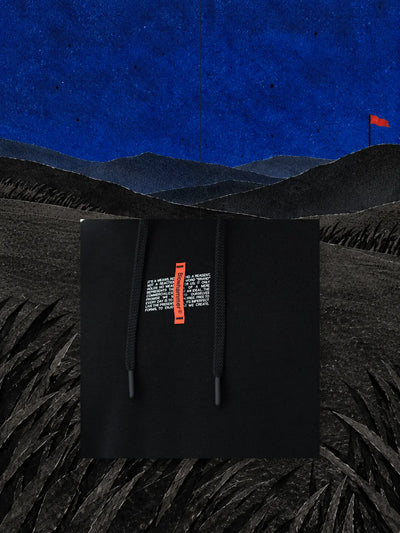 A piece of the chest of the black Flag hoodie, with the red label, hanging on a thread over a black paper hill under a blue sky.