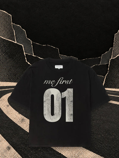The black boxy MeFirst01 t-shirt on a black paper track curving into the dark, beside a pale paper podium.