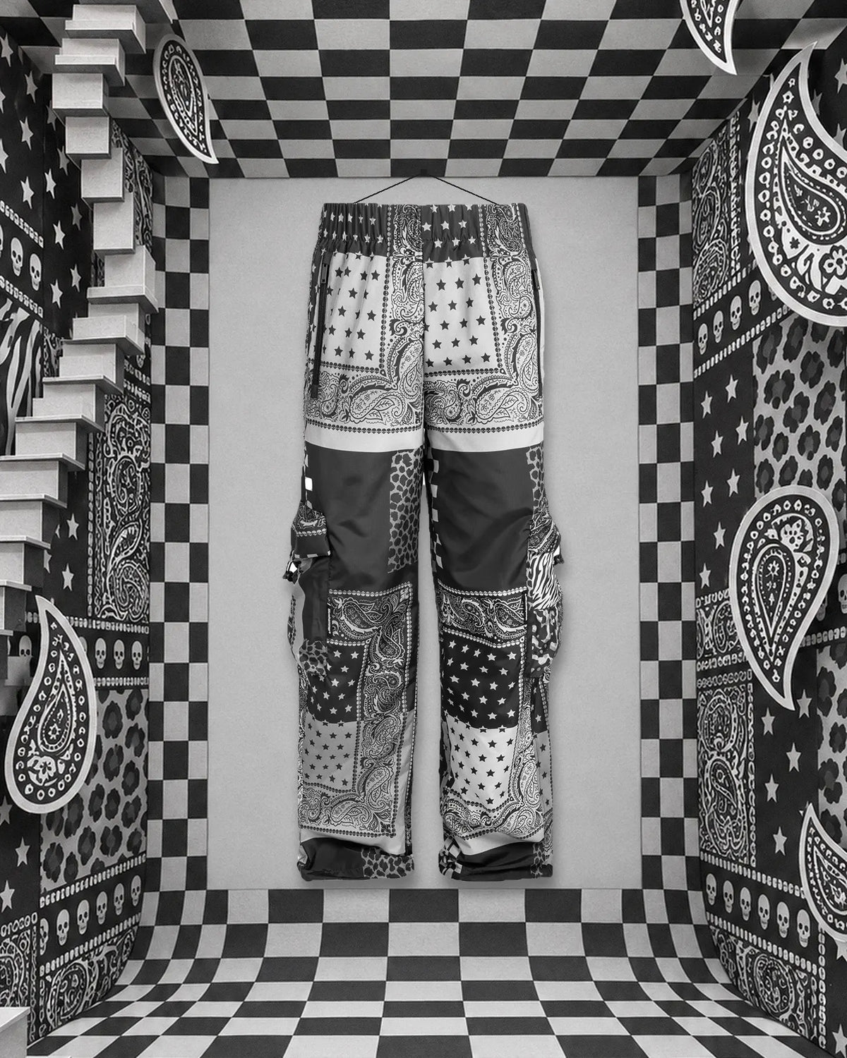El pantalón cargo Romanticbandana, patchwork de bandanas negras, blancas y grises, colgado con un hilo de un clavo en la pared del fondo de una habitación de papel de cuadros blancos y negros. A la izquierda una escalera blanca, en las paredes paisley, estrellas, rayas de cebra, manchas de leopardo y pequeñas calaveras.