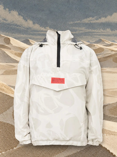 L'anorak avorio Strand Ivory fra dune di carta color sabbia, sotto un cielo di carta chiara.
