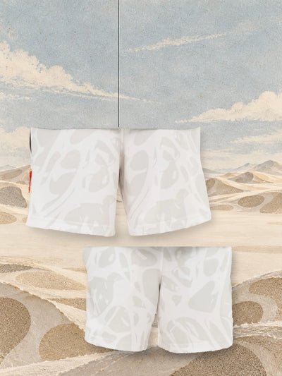 The ivory Strand Ivory bermuda shorts seen from the back, hanging on a thread, and from the front, above sand coloured paper dunes.