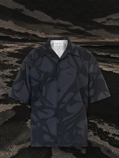 The black Strand short sleeve shirt on the bank of a black paper river, under a dark sky.