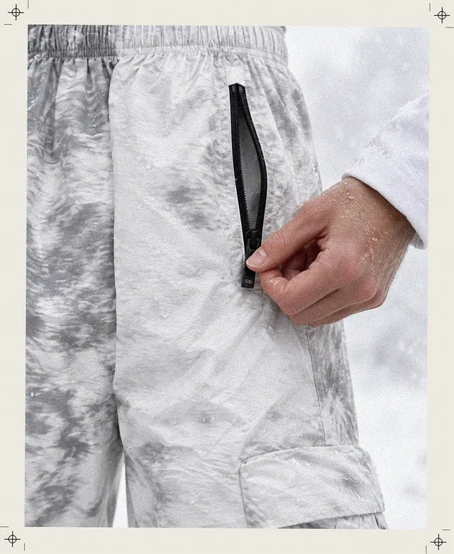 Una mano abre la cremallera negra del bolsillo del pantalón cargo Grey Noise, tejido blanco arrugado con nubes grises, con copos de nieve que se derriten sobre el tejido.