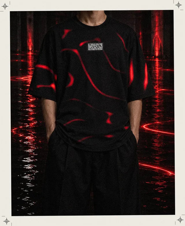 Uma pessoa fictícia, enquadrada do pescoço para baixo, veste a t-shirt Tide preta com os rastos vermelhos e está parada numa sala escura inundada, onde linhas de luz vermelha deslizam sobre a água preta.